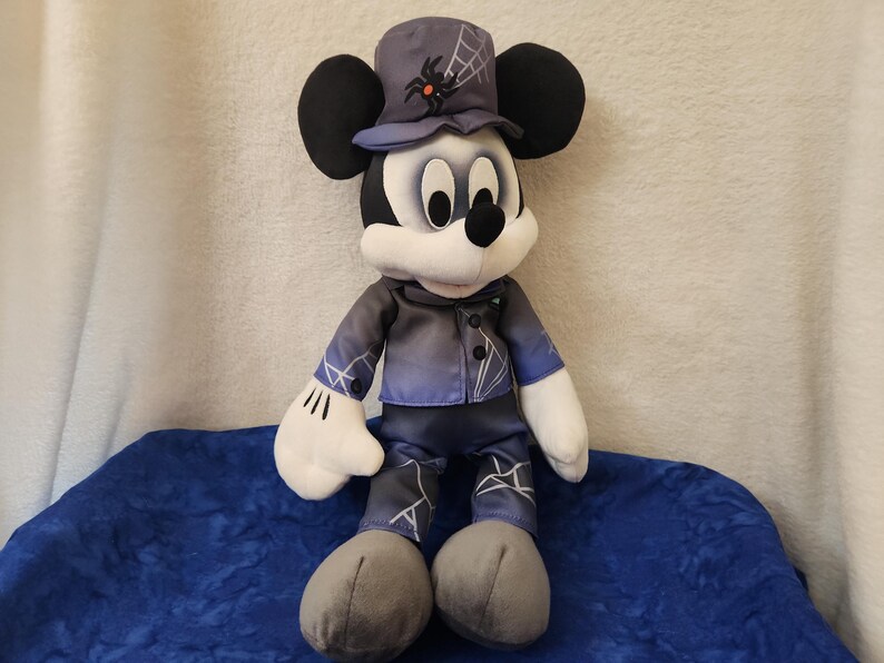 May include: A plush Mickey Mouse doll dressed in a Halloween costume. Mickey wears a dark blue jacket and pants with white accents, a matching hat with a spiderweb design, and gray shoes. The doll has black ears, a white face, and white gloves.