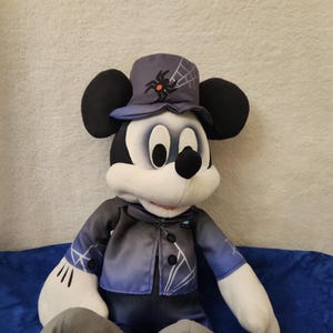 May include: A plush Mickey Mouse doll dressed in a Halloween costume. The doll wears a dark blue top hat with a spider and web design, a matching jacket, and black pants. The doll has black ears, a white face, and a black nose.