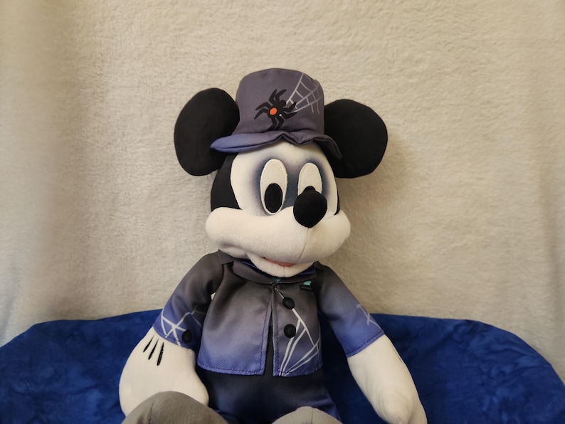 May include: A plush Mickey Mouse doll dressed in a Halloween costume. The doll wears a dark blue top hat with a spider and web design, a matching jacket, and black pants. The doll has black ears, a white face, and a black nose.