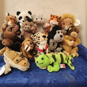 May include: A collection of plush animal toys, including a panda, tiger, lion, and crocodile. The stuffed animals are various sizes and colors, with tags attached. They are arranged on a blue fabric surface.