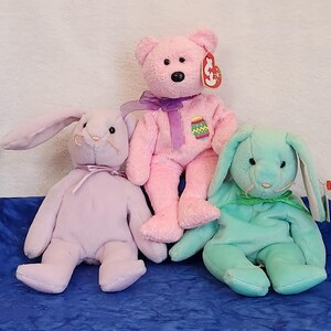 May include: Three plush toys: a pink bear with a purple ribbon and a tag that says "Beanie Babies", a light purple bunny with long ears, and a light blue bunny with long ears.