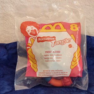 May include: A sealed clear plastic bag containing a Nickelodeon Tangle Twist-A-Zoid toy. The packaging features a red-haired clown graphic, the number 8, and the text Nickelodeon Tangle and Twist-A-Zoid. A curved orange and blue plastic toy is visible.