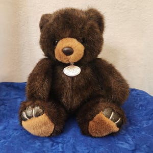 May include: A plush brown teddy bear with a light brown snout and paws. The bear has a silver pendant around its neck. The bear is sitting on a blue surface.