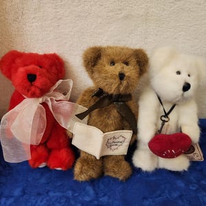 May include: Three plush teddy bears in red, brown, and white. The red bear has a white ribbon, the brown bear holds a small book, and the white bear has a red heart. The bears are on a blue surface.