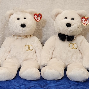 May include: Two white plush teddy bears, one with a black bow tie and the other with a floral collar. Both bears have black eyes, noses, and gold wedding ring accents. Each bear has a red heart-shaped tag.