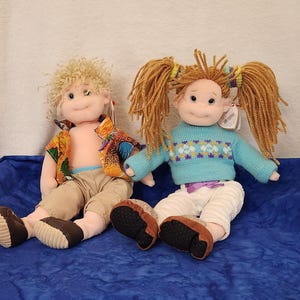 May include: Two plush dolls are seated on a blue surface. One doll has blonde curly hair, tan pants, and a colorful shirt. The other doll has pigtails, a blue sweater with a pattern, and white pants. Both dolls have brown shoes.