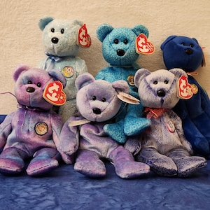 May include: A collection of six plush teddy bears in various shades of blue and purple. Each bear has a distinct tag and some have ribbons or bows. The bears are arranged on a blue fabric surface.
