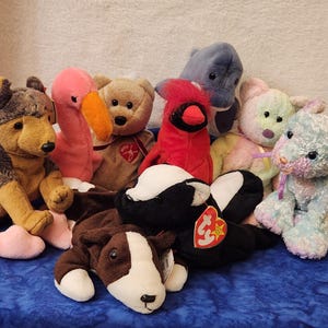 May include: A collection of plush stuffed animals, including a German Shepherd, flamingo, bear, cardinal, dolphin, bunny, lamb, skunk, and cow. The toys are in various colors and sizes, with tags attached.