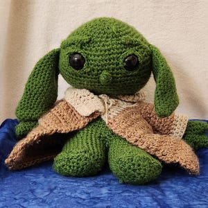 K&ouml;nnte beinhalten: Eine handgefertigte gr&uuml;ne H&auml;kelpuppe mit gro&szlig;en schwarzen Augen und langen Ohren. Die Puppe tr&auml;gt eine beige-braune H&auml;kelrobe. Die Puppe sitzt auf einer blauen Oberfl&auml;che.