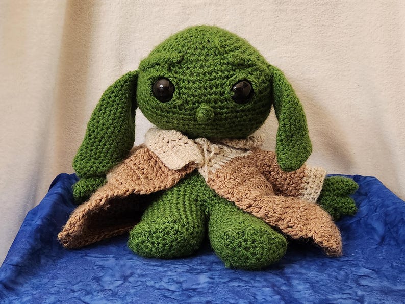 K&ouml;nnte beinhalten: Eine handgefertigte gr&uuml;ne H&auml;kelpuppe mit gro&szlig;en schwarzen Augen und langen Ohren. Die Puppe tr&auml;gt eine beige-braune H&auml;kelrobe. Die Puppe sitzt auf einer blauen Oberfl&auml;che.