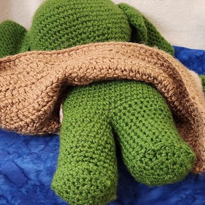 K&ouml;nnte beinhalten: Eine handgefertigte H&auml;kelfroschpuppe, haupts&auml;chlich gr&uuml;n, mit einer braunen Decke dar&uuml;ber. Die Beine des Frosches sind sichtbar, und die H&auml;kelarbeit hat ein strukturiertes Aussehen. Der Hintergrund ist ein blauer Stoff.
