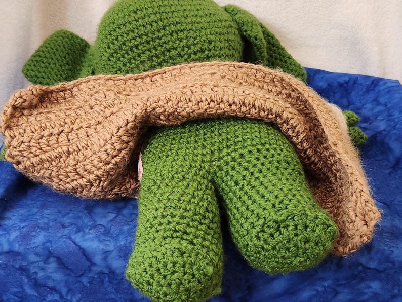 K&ouml;nnte beinhalten: Eine handgefertigte H&auml;kelfroschpuppe, haupts&auml;chlich gr&uuml;n, mit einer braunen Decke dar&uuml;ber. Die Beine des Frosches sind sichtbar, und die H&auml;kelarbeit hat ein strukturiertes Aussehen. Der Hintergrund ist ein blauer Stoff.