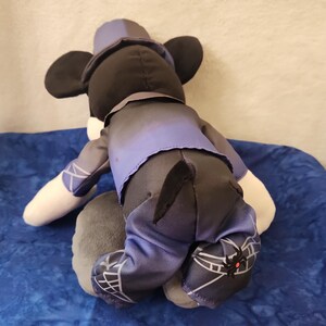 May include: A plush Mickey Mouse doll, viewed from the back, wearing a purple and black outfit. The doll has black ears, a matching hat, and white gloves. The outfit includes a vest, pants, and shoes with a spiderweb design.