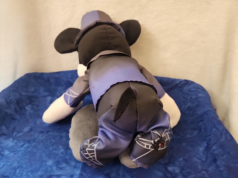 May include: A plush Mickey Mouse doll, viewed from the back, wearing a purple and black outfit. The doll has black ears, a matching hat, and white gloves. The outfit includes a vest, pants, and shoes with a spiderweb design.