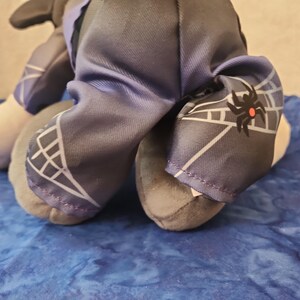 May include: A plush toy with gray and white feet and legs. The toy's clothing includes dark blue pants with a white web design and a black spider graphic. The toy is on a blue surface.