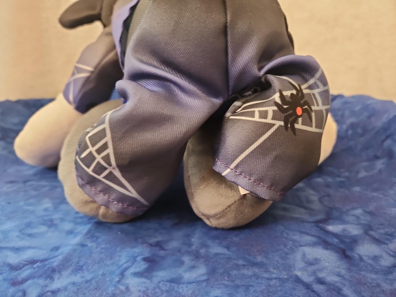 May include: A plush toy with gray and white feet and legs. The toy's clothing includes dark blue pants with a white web design and a black spider graphic. The toy is on a blue surface.
