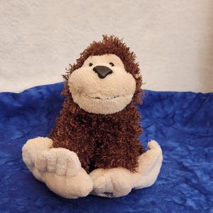 May include: A plush monkey toy featuring dark brown shaggy fur on its body and head, contrasted by a smooth, light beige face, hands, and feet. The stuffed animal has a black nose and a stitched mouth, sitting in an upright position.