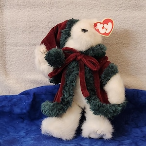 May include: A white plush bear wearing a burgundy and green Christmas outfit. The bear has a burgundy hat and vest with green trim. A red heart-shaped tag is attached to the bear's ear. The bear is on a blue surface.