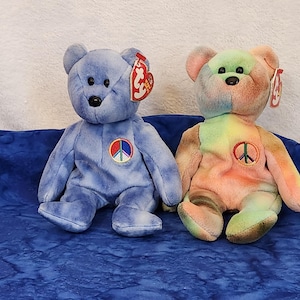 May include: Two tie-dye beanie baby bears with peace sign patches. The bear on the left is blue and the bear on the right is multi-colored.