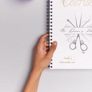 Puede incluir: Una libreta blanca de espiral con la palabra "Costura" escrita en letras doradas. La libreta tiene una ilustración gris de tijeras y agujas con el texto "Mis Diseños y Patrones" y "Creaciones y Confecciones 2021".