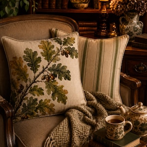 May include: Two square throw pillows featuring a beige fabric base. One pillow displays a detailed botanical print of green and yellow oak leaves with brown acorns on a branch. The second pillow features a pattern of vertical olive green stripes.