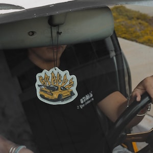 May include: A yellow car air freshener with the words "STANDS" and "2 Your Standards" printed on it. The air freshener is hanging from the rearview mirror of a car.