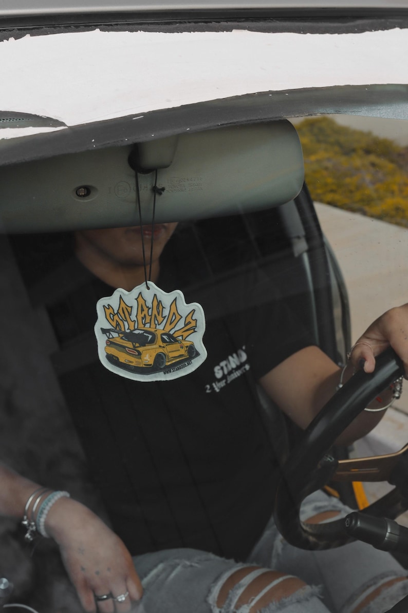 May include: A yellow car air freshener with the words "STANDS" and "2 Your Standards" printed on it. The air freshener is hanging from the rearview mirror of a car.