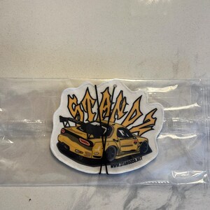 May include: Yellow car air freshener with the word "STANDS" in a stylized font. The car is a yellow Mazda RX-7 with a black rear wing. The air freshener has a white background and the text "www.standzco.net" is printed at the bottom.