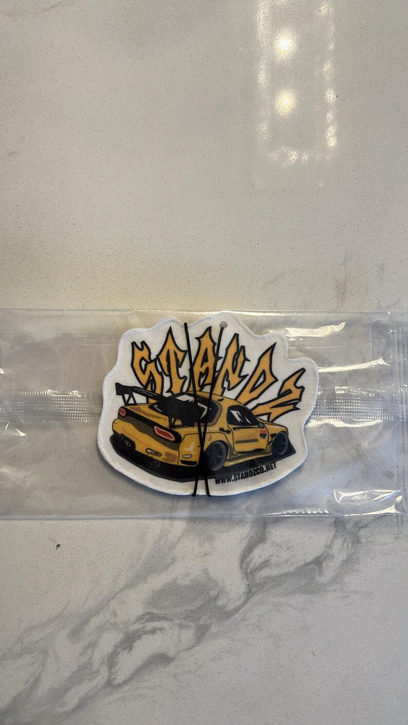May include: Yellow car air freshener with the word "STANDS" in a stylized font. The car is a yellow Mazda RX-7 with a black rear wing. The air freshener has a white background and the text "www.standzco.net" is printed at the bottom.