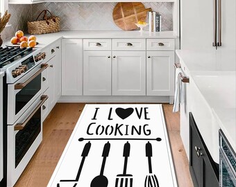 May include: White rectangular kitchen mat with the text "I LOVE COOKING" in black. Below the text are black silhouettes of kitchen utensils: a ladle, a spoon, a spatula, and a whisk. The mat is on a hardwood floor in a white kitchen.
