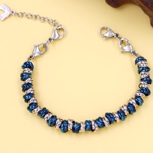 May include: A silver-toned bracelet with a series of dark blue and silver-toned beads. The bracelet has a chain extension and a lobster clasp closure. The bracelet is on a yellow surface with a dark brown wooden accent in the background.