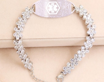 Könnte beinhalten: Ein silberfarbenes medizinische ID-Armband. Das Armband hat ein Blumendesign mit klaren Kristallen. Die zentrale ID-Platte hat ein medizinisches Symbol und Text. Das Armband liegt auf einer hellbeigen Oberfläche.