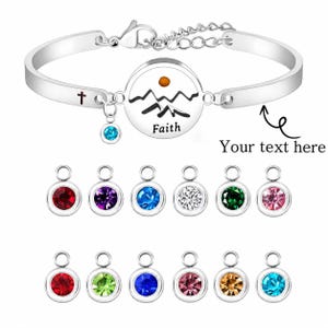 May include: A silver-toned metal bangle bracelet featuring a central circular charm with a mountain and sun graphic above the word Faith, a small cross symbol on the band, and a detachable charm with a blue crystal, plus twelve additional charms.