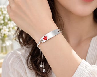 Op de afbeelding: Een zilverkleurige medische alarmarmband met een gladde, gebogen metalen identificatieplaat met een rood Star of Life-embleem. De armband heeft een fijne ketting met twee bungelende bedels, elk versierd met een kleine, heldere kristal.