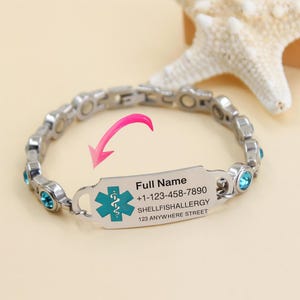 May include: A silver-toned medical ID bracelet with blue gemstone accents. The rectangular ID tag displays the medical symbol, contact information, and the text "Full Name", "Shellfish Allergy", and an address.