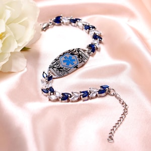 May include: Silver-toned medical ID bracelet with blue and clear gemstone accents. The central oval plate features a blue medical symbol and decorative black filigree. The bracelet is set against a pink satin background.