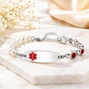May include: A medical alert bracelet featuring a central silver-toned metal plate with a red Star of Life emblem. One side of the bracelet consists of white pearls, while the other side features silver-toned links set with round red crystals.