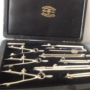 Könnte beinhalten: Ein offener schwarzer Koffer mit einem Satz silberner Zeichenwerkzeuge. Der Koffer enthält Zirkel, Teiler und andere Instrumente. Auf dem Koffer steht in Gold "A.G. Thornton Ltd. Manchester".