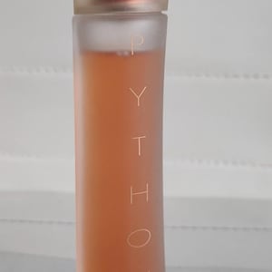 Puede incluir: Una botella de perfume alta y cilíndrica con una tapa transparente esmerilada y un líquido color melocotón en su interior. La palabra "PYTHON" está impresa verticalmente en la botella. La botella tiene un diseño minimalista.