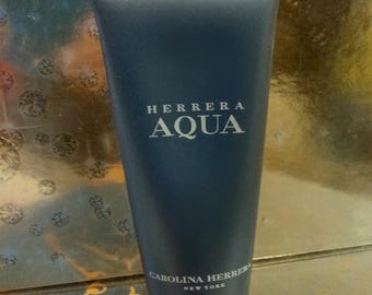 Può includere: Un tubetto di balsamo dopobarba blu scuro con la scritta "HERRERA AQUA" e "CAROLINA HERRERA NEW YORK" in bianco. Il prodotto è su una superficie riflettente.