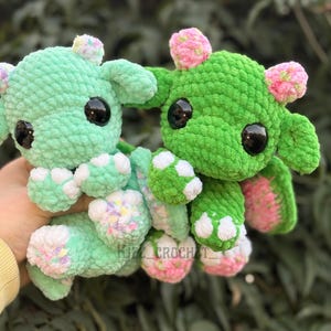 May include: Two crocheted plush toys in shades of green, with large black eyes and small pink and white accents. The toys have rounded bodies and are in a playful pose. The text "KIZZ_CROCHET_" is visible.