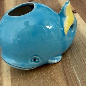 May include: A glossy ceramic whale figurine in a bright blue color with a yellow tail fin. The whale has a rounded body, a small opening on the top, and a smiling face with a single visible dark eye. The surface of the ceramic is smooth and reflective.