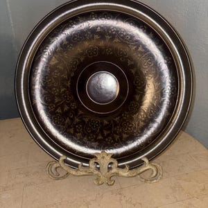 May include: A decorative, round, dark brown tray with a floral pattern and a silver centre. The tray is supported by a cream-coloured, ornate stand. The tray's rim is a lighter shade of brown.