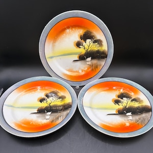 May include: Three white porcelain plates with a hand-painted design of a lake scene with a tree and birds. The plates have a blue and orange rim.