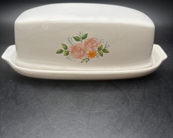 Könnte beinhalten: Weißer Keramik-Butterbehälter mit Deckel. Die Schale hat ein Blumendesign mit rosa Rosen und orangefarbenen Blüten. Der Boden hat einen leicht erhöhten Rand mit kleinen Griffen auf jeder Seite. Der Butterbehälter ist für eine Standardbuttergröße ausgelegt.