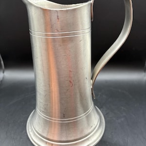 May include: A silver pewter jug with a rounded spout and a handle. The jug has a slightly flared base and a smooth, polished finish.