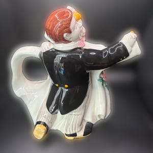 May include: A ceramic teapot depicting a dancing couple, the groom in a black tuxedo and the bride in a white dress. The teapot has a white handle and spout, with the couple's faces and hair detailed in color.