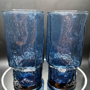 May include: Two blue textured drinking glasses. The glasses have a unique, organic texture and a dark blue color. The base of the glasses is a darker shade of blue. The glasses are sitting on a reflective surface.