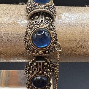 May include: A silver bracelet with three blue glass stones set in ornate silver settings. The bracelet has a hinged clasp and a small dangling chain.