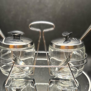 Peut inclure: Un ensemble sucrier et crémier vintage. L'ensemble comprend deux pots en verre transparent avec des couvercles argentés et de petites cuillères. Les pots sont maintenus dans un support en métal argenté avec une poignée.